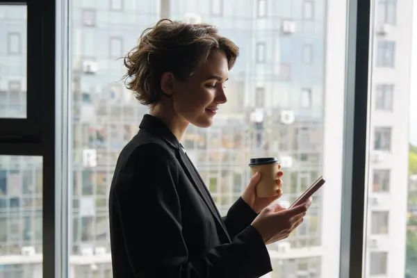 Portrait of corporate woman reading message on mobile phone, drinking coffee, standing near office mirror and smiling.