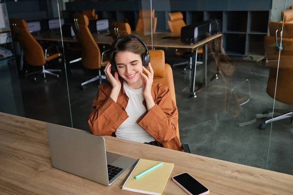 Portrait of working woman, social media manager in office, sitting at workplace, connects to online meeting, video chat on headphones, looking at laptop.