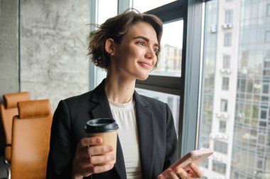 Başarılı kadın girişimcinin portresi ofiste bir fincan kahve içer, akıllı telefon tutar, konferans odasında müşteriyi bekler..