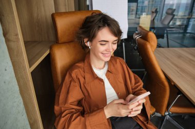 Kadın çalışanın portresini kapat, günlük giysiler içindeki girişimci, ofiste çalışıyor, akıllı telefon kullanıyor, kablosuz kulaklıkla müzik dinliyor..