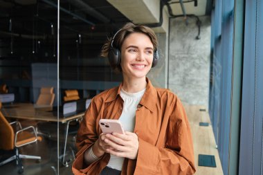 Dijital göçebe portresi, kablosuz kulaklıklı kadın yönetici, pencerenin yanında duruyor, cep telefonu tutuyor..