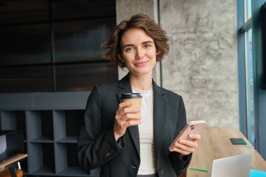 Başarılı bir iş kadınının portresi, sabah kahvesi, ofis penceresinin yanında duruyor ve gülümsüyor, cep telefonu uygulamasından haberleri okuyor..