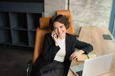 Ofisinde oturan, telefonlara cevap veren, akıllı telefonunu ve laptopunu kullanan, pencerenin yanında çalışan genç bir iş kadınının portresi..