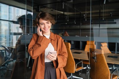 Dijital göçebe portresi, ofiste yönetici olarak çalışan kadın, kablosuz kulaklıklar ve akıllı telefonlarla ofiste duran, birini arayan ve kulaklıkla konuşan..
