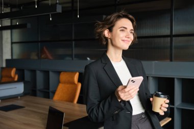 Genç ve başarılı bir iş kadınının konferans odasında, akıllı telefonlu bir ofiste durup kahve içerken camdan dışarı bakıp gülümsemesi..