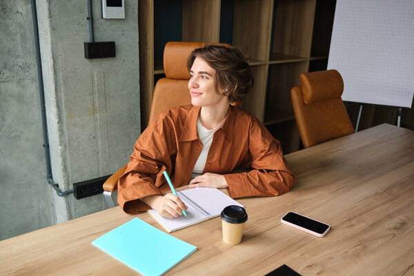 Portrait of young female entrepreneur, employee working in office, writing down, making notes in conference room, smiling.