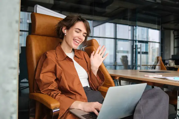 Portrait of professional woman says hello, waves hand at laptop screen, sits in office, works with team online, connects to meeting on computer.