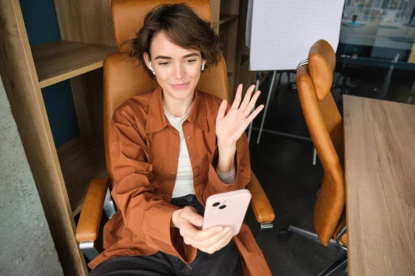 Portrait of woman saying hello, talking to smartphone, video chatting, having an online meeting in her office, using mobile phone for conversation.