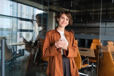 Gülümseyen güzel bir kadının portresi, kablosuz kulaklık takan bir menajer, elinde akıllı telefon, ofiste çalışıyor..