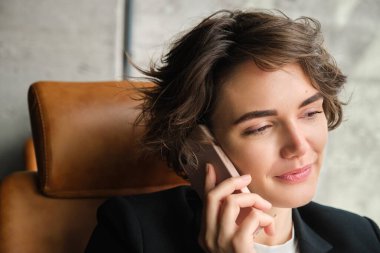 Gülümseyen esmer kadın, cep telefonuyla konuşan iş kadını, telefonla konuşan ve gülümseyen deri koltukta oturan,.