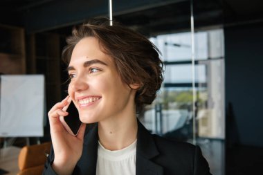 Başarılı bir iş kadınının cam duvarlı bir ofiste telefon konuşması yapması, gülümsemesi ve pazarlıklardan memnun görünmesi..