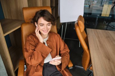 Ofiste çalışan bir kadının portresi, akıllı telefon kullanan, molasında sandalyede oturan ve gülümseyen bir çalışan..