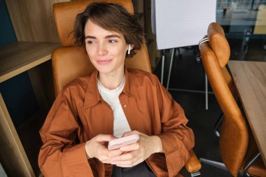 Ofiste çalışan bir kadının portresi, akıllı telefon kullanan, molasında sandalyede oturan ve gülümseyen bir çalışan..