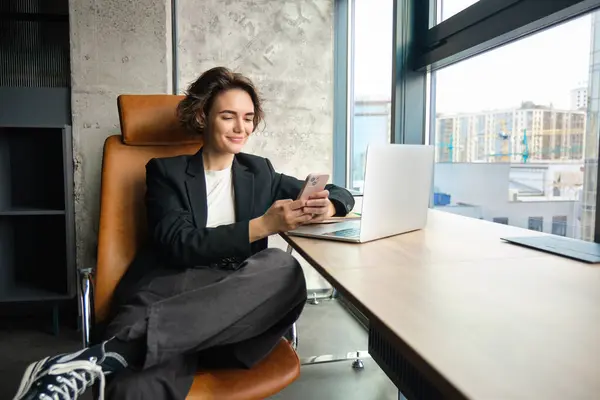 Portrait of corporate woman, saleswoman in an office, using smartphone and laptop, smiling while texting message on mobile phone.