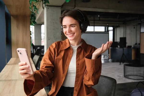 Portrait of beautiful smiling woman, company employee, sitting in office, talking on smartphone, wearing headphones, video chats, connects to online meeting on mobile phone.