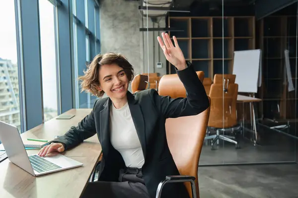 Portrait of woman working on laptop, businesswoman saying hello, waving at co-worker in her office and smiling, saying hello to colleague.