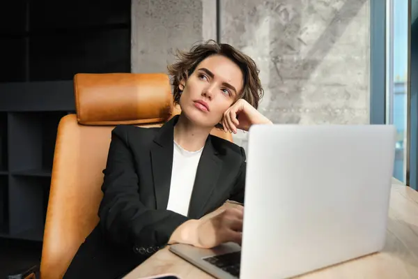 Portrait of woman focused on work, corporate lady manager in suit, sitting in office with laptop, thinking, looking up with thoughtful face expression.