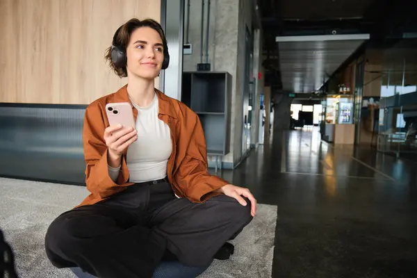 Image of stylish young company employee, sitting in office on floor, resting in lounge room at co-working space, holding mobile phone, wearing headphones.