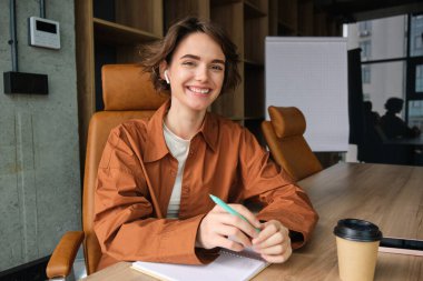 Başlangıç projesinde çalışan, notlar alan, elinde kalem tutan, kağıt okuyan, ofiste oturan neşeli genç bir kadının portresi..