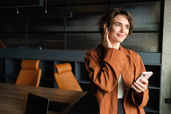 Portrait of woman, company employee in casual clothes standing in office with mobile phone, talking to client with wireless headphones and smiling.
