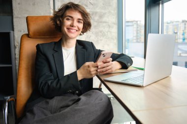 Ofisinde çalışan kurumsal bir kadının portresi, akıllı telefondaki mesajlara cevap vermek, pencerenin önünde dizüstü bilgisayarla oturmak, takım elbise giymek..