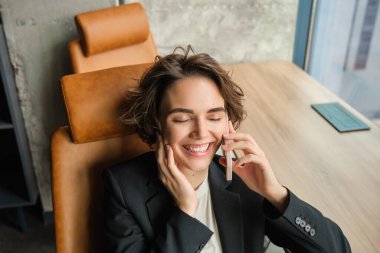 Takım elbiseli bir iş kadını portresi, ofisinde oturuyor ve telefonuna mutlu bir gülümsemeyle cevap veriyor, telefonda pazarlık yapıyor..