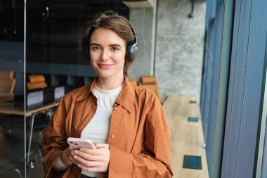 Ofisteki genç modern kadın portresi, kablosuz kulaklıklı dijital göçebe, iş yerinde akıllı telefonuyla duruyor ve gülümsüyor, pencerenin yanında poz veriyor..