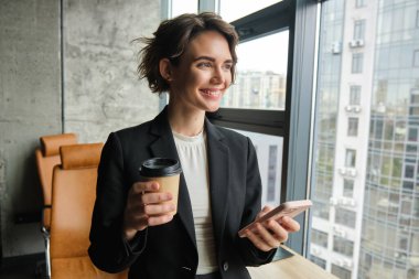 Kadın girişimci portresi, iş yerinde dikilip elinde akıllı telefon, pencerenin yanında kahve içiyor..