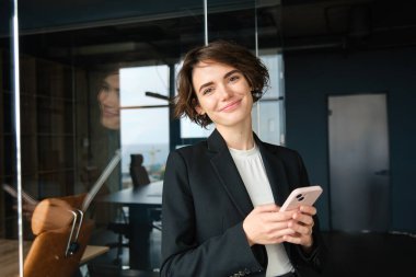 Siyah takım elbiseli şık bir iş kadını, akıllı telefonlu cam duvarın yanında duruyor, ofiste çalışıyor..