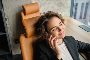 Ofisinde oturan, telefonlara cevap veren, akıllı telefonunu ve laptopunu kullanan, pencerenin yanında çalışan genç bir iş kadınının portresi..