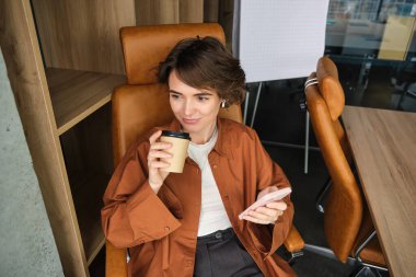 Ofiste kahve içen, akıllı telefondan sosyal medyaya kayan, elinde cep telefonu olan bir kadının portresi..