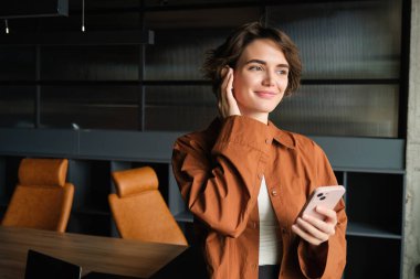 Gülümseyen genç kadın, iş adamı, ofis konferans odasında duruyor, kablosuz kulaklıklarla telefon görüşmesine bağlanıyor ve dışarıdan bakıyor, şirket için çalışıyor..