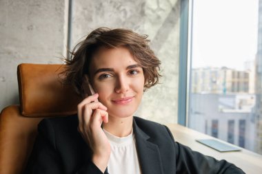 Şirket insanları ve iş kavramı. Takım elbiseli genç bir kadın, ofiste oturuyor ve sohbet ediyor, telefona cevap veriyor ve gülümsüyor, müşteriyle konuşuyor..