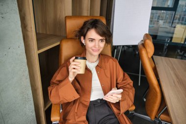 Ofiste kahve içen, akıllı telefondan sosyal medyaya kayan, elinde cep telefonu olan bir kadının portresi..