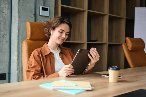 Portrait of young woman CEO, businesswoman in casual clothes, sitting in her office with digital tablet and cup of coffee, working on project, reading.