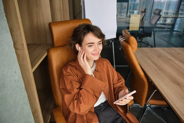 Top view of young woman working in office, co-working space, sits in her chair with wireless headphones and mobile phone, listens to music, calls someone with earphones.