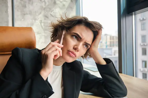 Close up portrait of working woman in an office, having difficult conversation, frowning while answering phone call, sitting in suit.