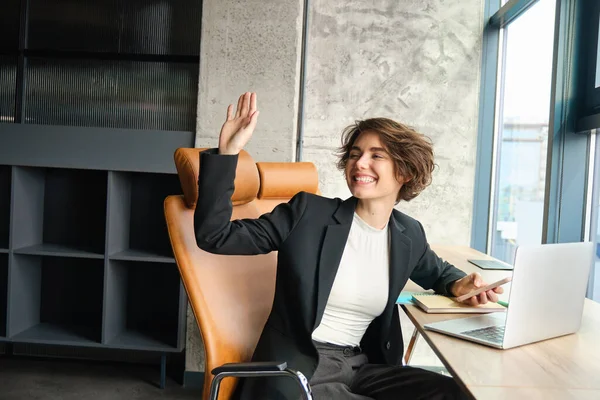 Portrait of young successful woman, corporate lady in an office, turn around and saying hello, waving hand to say hi to colleague, sitting with laptop and mobile phone.