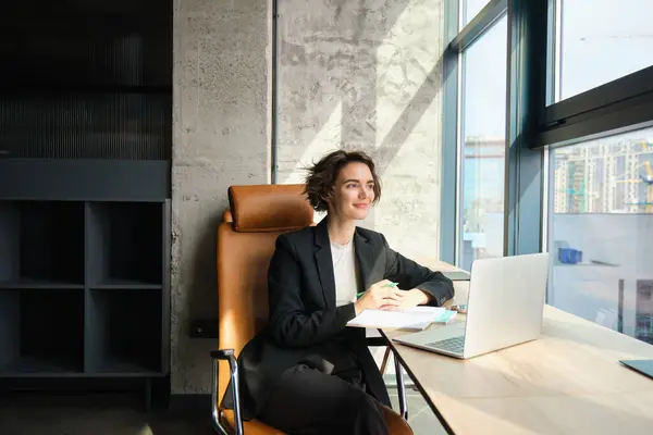 Portrait of young company employee, manager sitting in suit in her office, working on laptop, waiting for business meeting, looking outside window with confidence.
