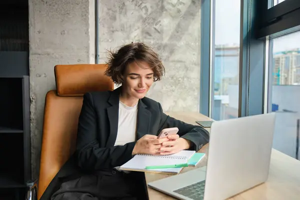 Portrait of corporate woman in suit, sits in office, messaging someone, replying to friend on smartphone, working with laptop, having lunch break.