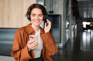 Mutlu genç bir kadının portresi, ofiste yönetici, dinlenme odasında öğle yemeğinde dinleniyor, akıllı telefonu tutuyor, kablosuz kulaklıkla müzik dinliyor, ortak çalışma alanını kullanıyor..