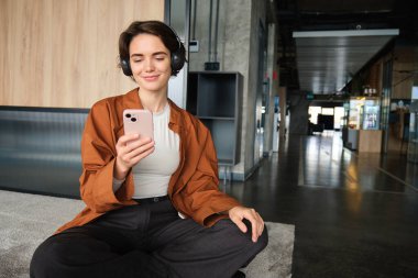 Kadın çalışanın portresi, ofiste modern yönetici, dinlenme odasında yerde oturuyor, akıllı telefon kullanıyor, cep telefonuna bakıyor ve gülümsüyor..