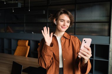 İş görüşmesine bağlanan kadın görüntüsü, cep telefonuyla görüntülü konuşma, akıllı telefon uygulamasına el sallama ve gülümseme, sıradan kıyafetlerle bir ofiste duruyor..
