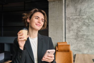 Başarılı bir iş kadınının portresi, sabah kahvesi, ofis penceresinin yanında duruyor ve gülümsüyor, cep telefonu uygulamasından haberleri okuyor..