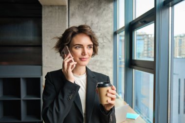 Ofiste kahve içen esmer kadının portresi. Takım elbiseli genç bir iş kadını birlikte çalışma alanındaki pencereyi temizler, müşteriyi arar, telefonda sohbet eder..