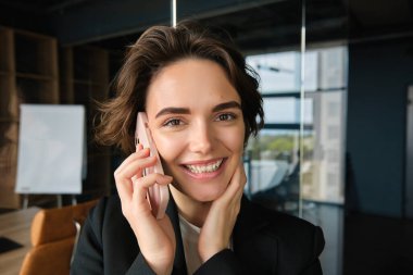 Gülümseyen satıcının yakın portresi, ofis müdürü konferans odasında duruyor, telefona cevap veriyor, cep telefonuyla biriyle konuşuyor, harika haberler alıyor ve seviniyor..