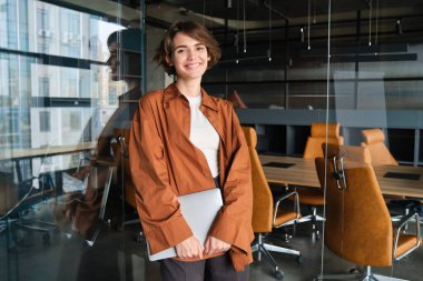 Genç kadın programcının portresi, ofiste çalışan dijital göçebe, dizüstü bilgisayarıyla duruyor ve gülümsüyor..