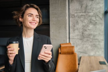 Başarılı bir iş kadınının portresi, sabah kahvesi, ofis penceresinin yanında duruyor ve gülümsüyor, cep telefonu uygulamasından haberleri okuyor..