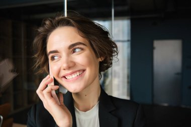 Başarılı bir iş kadınının cam duvarlı bir ofiste telefon konuşması yapması, gülümsemesi ve pazarlıklardan memnun görünmesi..