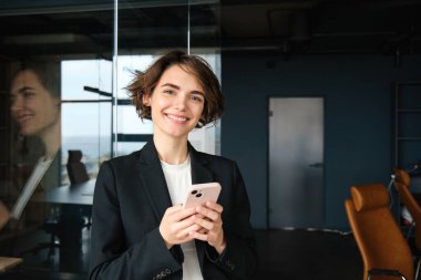 Takım elbiseli bir kadının portresi, ofis odasında akıllı telefonlu ve gülümseyen..